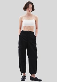 Bílé top s krátkým rukávem a tenkými ramínky, černé cargo kalhoty s elastickým pasem a kapsami, stylizované s černými mokasíny. Neutrální šedé pozadí.