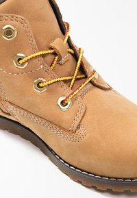 Botín de ante marrón con cordones amarillos y rojos, ojales dorados y costuras blancas. Cuenta con una suela gruesa para mayor tracción.