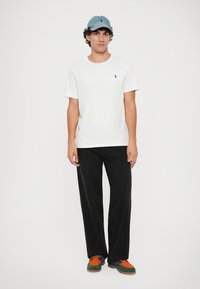 Camiseta branca com um logótipo azul, calças largas pretas e sapatos coloridos com uma mistura de toques laranja, verde e roxo. Boné de baseball em denim.