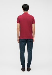 Homme debout de dos portant un polo rouge à manches courtes, un jean bleu foncé et des chaussures marron sur un fond clair uni.