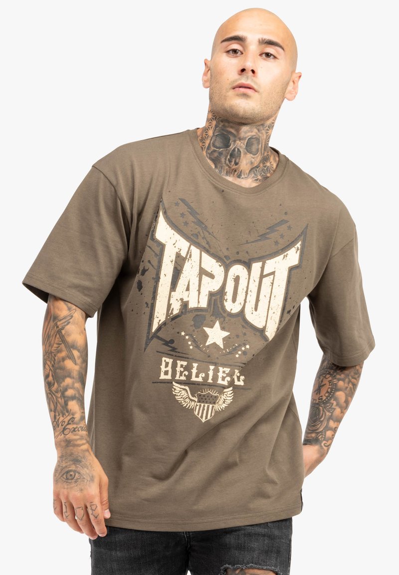 Olívazöld pamut póló, melyen középen nagy, elhalványult fehér grafika található "TAPOUT", csillag és szárny mintákkal. Rövid ujjú és laza fazonú.