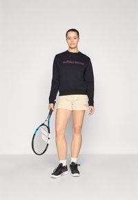 Sweatshirt preto com "BJÖRN BORG" bordado em rosa, combinado com calções beges. Segura uma raquete de ténis, vestindo sapatos pretos e brancos.