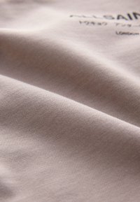 Detail mäkkej, svetlo béžovej tkaniny s jemnou textúrou a čiastočne viditeľným rozmazaným potlačeným textom navrchu.