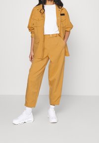 Senapsgul arbetsdress och matchande byxor i avslappnad passform, med en vit t-shirt under och vita sneakers. Utrustad med fickor.