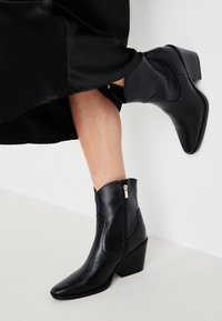Botines negros de cuero liso, con puntera afilada, detalle de costura en ángulo y cremallera lateral con un tirador dorado.
