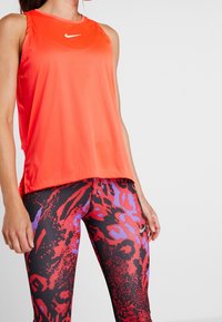 Femme portant un haut sans manches orange vif et des leggings noirs avec des motifs abstraits rouges et violets, debout devant un fond blanc.