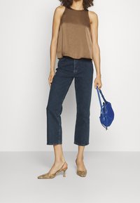 Top sin mangas de satén marrón, jeans cropped de mezclilla oscura, tacones de slingback dorados con patrón y un bolso pequeño azul.