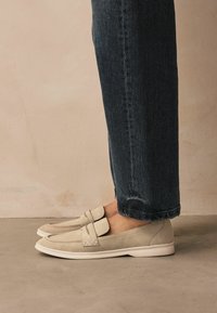Bežid suede loafers valge kummitaldadega, kahe dekoratiivse rihma ja tikanditDetailidega. Kaasnevad tumedat teksapüksid.