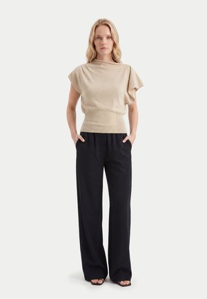 Frau steht mit Händen in den Taschen, trägt beige kurzärmliges Oberteil mit drapiertem Schulterbereich und schwarze weite Hosen vor weißem Hintergrund.