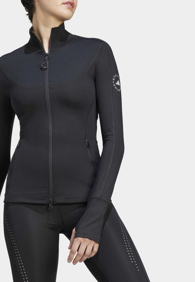 Chaqueta deportiva negra con cremallera fabricada en material elástico, con mangas largas, orificios para los pulgares y un logo en la manga izquierda.