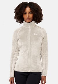 Hellbeige Fleecejacke mit hohem Kragen, Reißverschluss und Seitentaschen. Der Stoff hat eine weiche Textur und zeigt ein Logo auf der Brust.