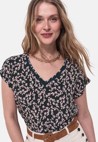 Blusa floreale a maniche corte di colore verde scuro con motivi rosa, scollatura a V e bordo in pizzo; abbinata a una cintura di colore chiaro.