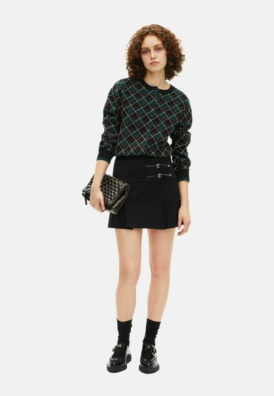 Mladá žena s kučeravými vlasmi, oblečená v tmavej károvej kardigáne, čiernej mini sukni s prackami, čiernych ponožkách a mokasínach, ktorá v ruke drží prešívanú kabelku.