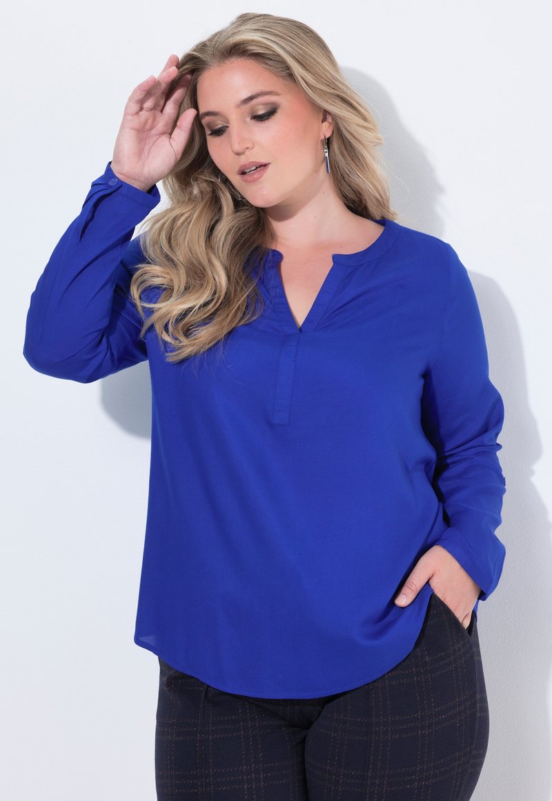 Blusa azzurra a maniche lunghe con scollatura a V e orlo curvo, realizzata in tessuto leggero e con dettagli di cucitura sottili. Abbinata a pantaloni scuri con fantasia.