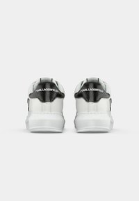 Ett par vita sneakers med svarta hälflikar som visar "KARL LAGERFELD"-logotyp, sett bakifrån mot en enkel bakgrund.
