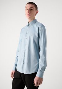Camisa de manga larga azul claro hecha de una tela suave, con un cuello abotonado, botones blancos y un sutil logo en el pecho.