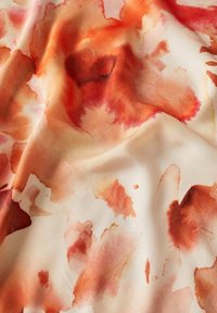 Tissu doux avec des motifs de taches aquarelles orange et rouges sur un fond beige clair, montrant de douces pliures et une texture délicate.