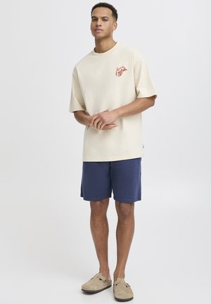 Beige oversized T-shirt met rood ontwerp, gecombineerd met marineblauwe korte broek. Model draagt beige instap schoenen. Eenvoudig en casual ontwerp, katoenen stof.