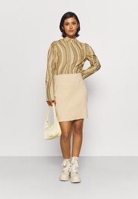 Top de manga longa bege com riscas abstratas castanhas, combinado com uma saia mini bege clara. A modelo segura uma mala de ombro creme, usando botas brancas.