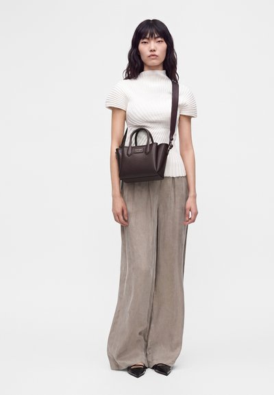 Femme portant un haut blanc côtelé à manches courtes, un pantalon large beige, des chaussures noires pointues et un sac bandoulière marron foncé.