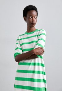 Abito bianco a t-shirt con strisce orizzontali verdi, caratterizzato da un collo rotondo e maniche a tre quarti. Tessuto morbido e testurizzato, vestibilità rilassata.
