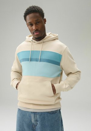 Junger Mann mit den Händen in den Taschen, trägt einen beigen Hoodie mit drei blauen Streifen quer über der Brust und blaue Jeans.