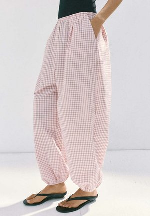 Pantalon ample à carreaux roses et blancs avec des poignets élastiques, porté avec des tongs noires, montré de la taille aux pieds avec une main dans la poche.