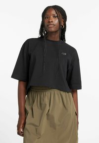 Camiseta negra recortada hecha de tela suave con un cuello redondo y mangas cortas, con un detalle sutil del logo. Combinada con una falda caqui.