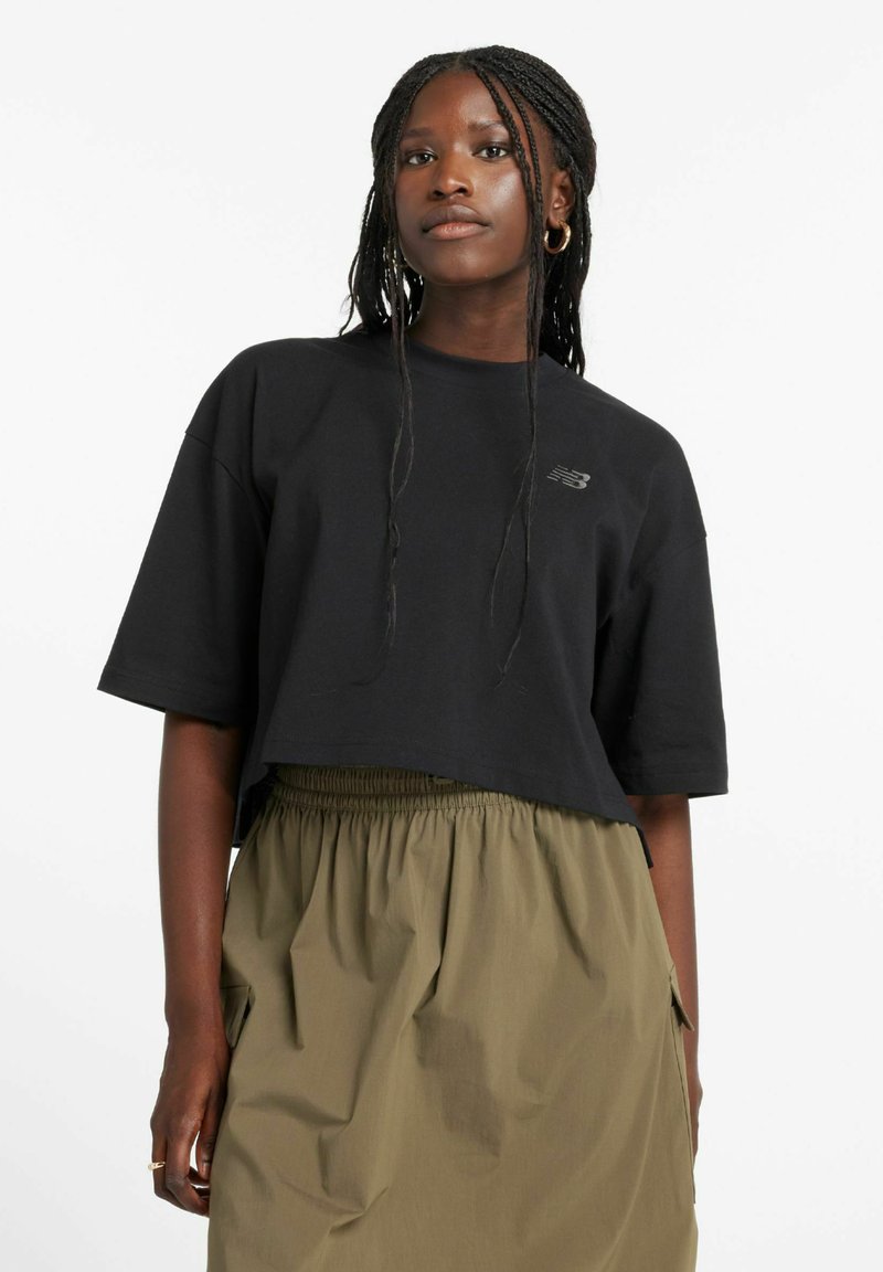 Camiseta negra recortada hecha de tela suave con un cuello redondo y mangas cortas, con un detalle sutil del logo. Combinada con una falda caqui.