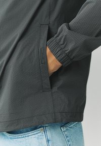 Jachetă în carouri alb-negru cu manșete elastice și buzunar lateral, purtată împreună cu blugi din denim de culoare albastru deschis. Material neted, ușor.