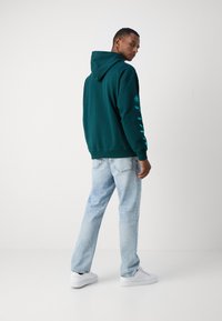 Felpa verde scuro con cappuccio, decorata con un design floreale turchese lungo la manica, abbinata a jeans azzurri chiari e sneaker bianche.