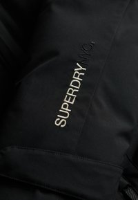 Fekete dzseki sima textúrával, az elején világosszürke hímzett "SUPERDRY NYC" felirattal, amely hangsúlyozza a márkát.