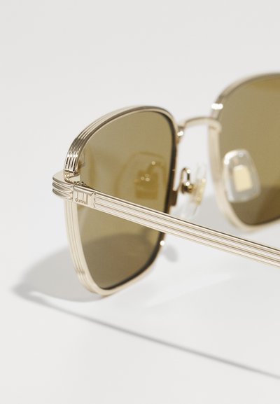 Lunettes de soleil en métal doré avec des verres teintés bruns, présentant un détail rainuré sur la monture et un accent de logo près de la charnière. Fini poli.