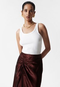 Weißes geripptes Tanktop, kombiniert mit einem glänzenden burgunderfarbenen Rock mit Raffung und Kordelzugdetails an der Taille. Akzente aus goldenem Schmuck.