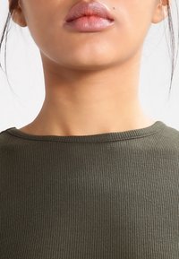 Olivengrøn ribbet top med bred halsudskæring og glat tekstur, med synlig syning omkring kraven og skuldrene.