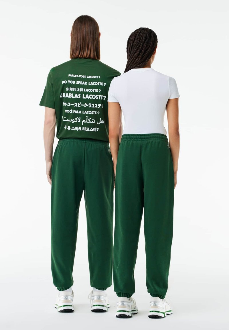 Zielona bawełniana koszulka z wielojęzycznym napisem na plecach; połączona z zielonymi dresami, obie z elastycznymi paskami w talii i luźnym krojem.
