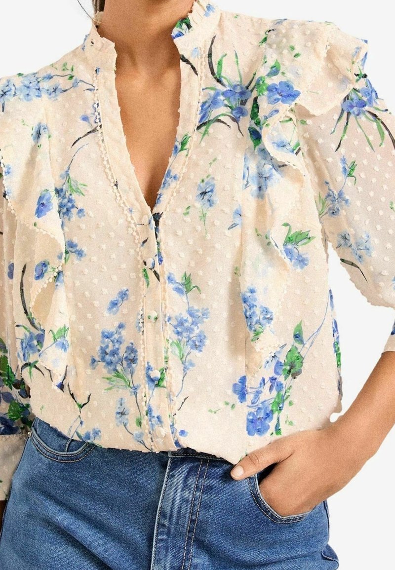 Florale Bluse aus durchscheinendem, cremefarbenem Stoff mit blauen und grünen Blumenmustern, texturiert mit Punkten, mit Rüschensäumen und einer Knopfleiste.