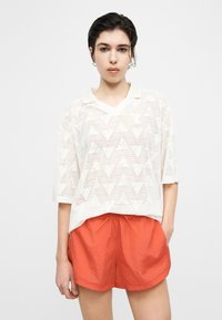 Γυναίκα με κοντά σκούρα μαλλιά που φοράει λευκό πουκάμισο με μοτίβο από διάτρητο ύφασμα και φωτεινά πορτοκαλί σορτς, στέκεται μπροστά σε ένα απλό λευκό φόντο.