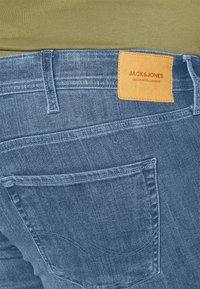 Blue denim jeans with a visible back pocket, featuring a rectangular tan leather patch labeled "JACK & JONES." Textured fabric with fading details.