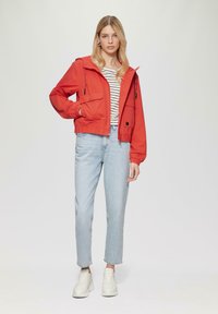 Rote Kapuzenjacke mit elastischen Bündchen und zwei Fronttaschen, kombiniert mit einem gestreiften Shirt und hellblauen geraden Jeans. Weiße Sneaker.