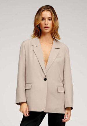 Bézs blézer beszabott hajtókával, egysoros gombolással és oldalzsebekkel. Simogatott anyagból készült, laza szabással és hosszú ujjakkal.