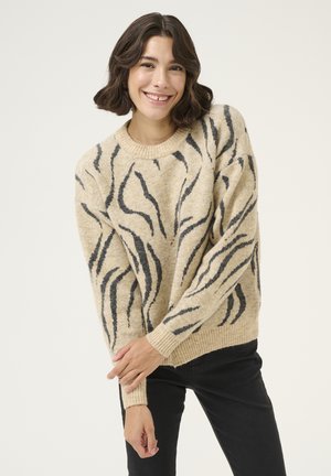 Pull beige avec un motif rayé noir en forme de tigre, col rond et poignets côtelés. Tissu tricoté doux et texturé. Porté avec un pantalon noir.
