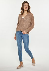Brede gebreide cardigan in lichtbruin met een V-hals en vijf knopen, voorzien van een gedetailleerd opengewerkt patroon. Gecombineerd met blauwe denim jeans en beige flats.