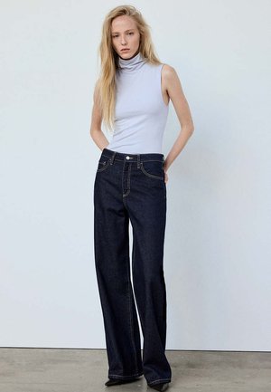 Blonde Frau trägt ein ärmelloses weißes Rollkragentop und dunkle weite Jeans und steht an einer schlichten Wand.