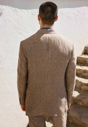 Mann mit kurzen Haaren steht vor einer weißen strukturierten Wand, trägt einen hellbraunen Nadelstreifenanzug, in der Nähe von steinernen Stufen im Freien.