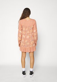 Kvinde med langt krøllet hår iført en pink blomstret langærmet kjole og sorte høj-top sneakers, som står med ryggen til mod en hvid baggrund.