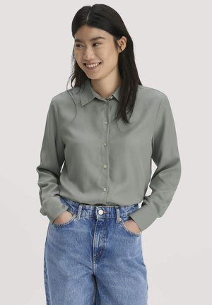 Frau mit langen dunklen Haaren, die ein graues Knopfhemd und blaue Jeans trägt, mit den Händen in den Taschen, lächelnd und seitlich schauend.
