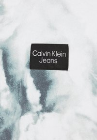 Calvin Klein Jeans-märke på tyg med ett ljust grått och vitt abstrakt mönster, med en svart rektangulär etikett som visar varumärket i vit text.