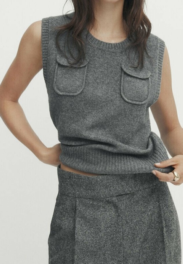 Femme portant un haut en maille gris sans manches avec des poches avant et une jupe grise taille haute assortie, tirant légèrement le haut vers le haut.