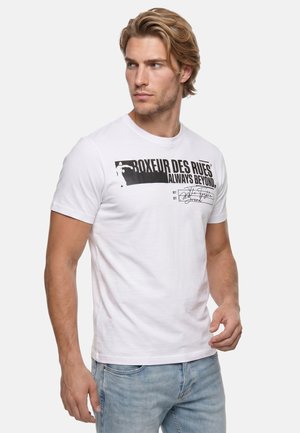 Mann mit blondem Haar, der ein weißes kurzärmeliges T-Shirt mit schwarzem Text „Boxeur Des Rues Always Beyond“ und hellblaue Jeans trägt, schaut nach links.